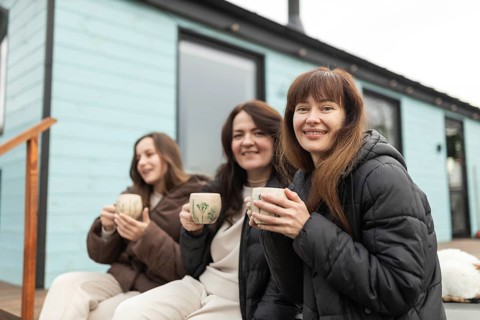Three female friends share a joyful moment while holding coffee mugs outside a modern home. They