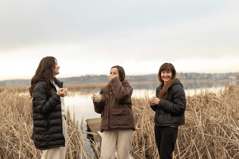 Three female friends share laughter and conversation while holding warm drinks, standing against a