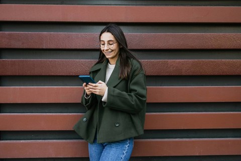 A woman in a green coat is smiling while using her smartphone She stands against a decorative