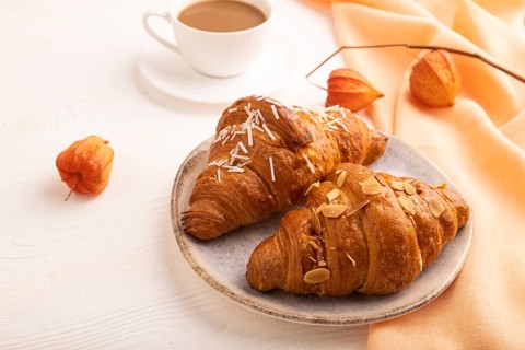 Croissant on blue plate on white wooden background and orange linen textile, cup of coffee, side