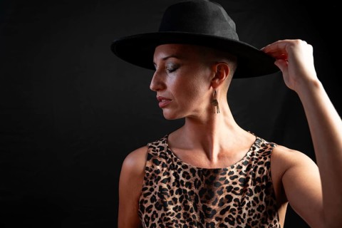 Portrait of a bald woman wearing a leopard print dress and black hat. She adjusts her hat,