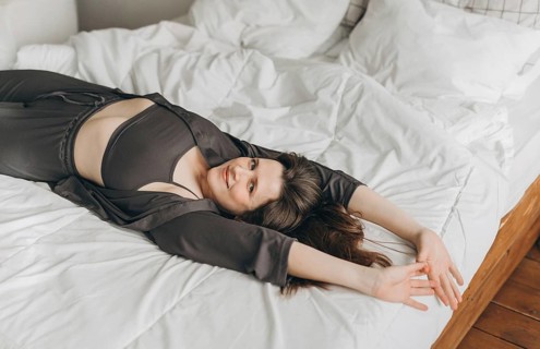 A cheerful young woman lies on a white bed with her arms stretched out, smiling and looking relaxed