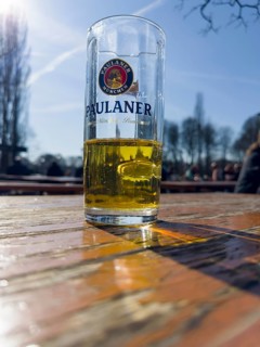 Half-filled Paulaner beer glass on a weathered beer garden table, behind it beer garden visitors,