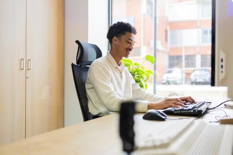 A professional works at a computer in a modern office setting, with natural light streaming through