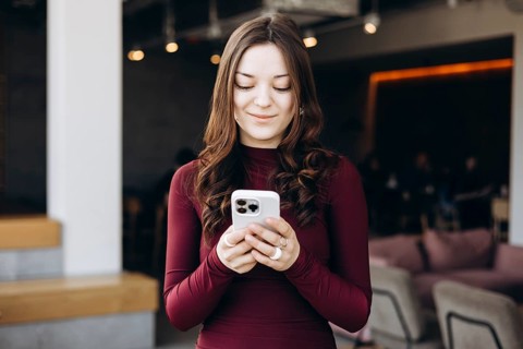 A young female student with long hair stands indoors, focused on her smartphone. She wears a