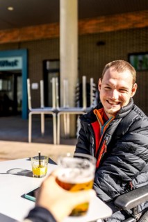 Young disabled man smiling and enjoying a beer with a friend at an outdoor cafe, promoting