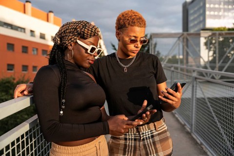 An LGBT multiethnic couple stands on a bridge, holding phones and enjoying each other's company.
