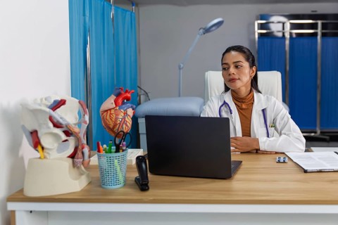 A female doctor in a white coat sits at her desk, focused on her laptop, with anatomical heart and