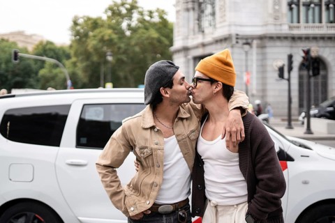 A Colombian gay couple shares a tender kiss on a city street, showcasing their affection. They wear