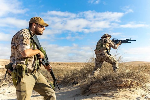 Two armed soldiers in desert terrain, wearing tactical gear and camouflage. Engaged in military