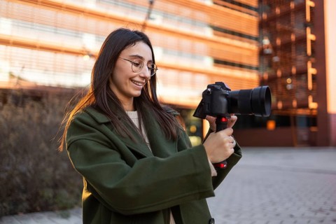 A woman wearing a green coat stands in an urban environment, holding a camera The warm light