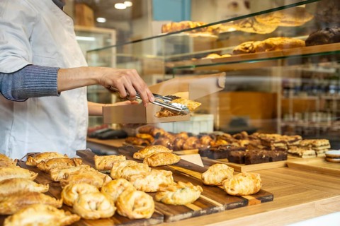 Baker's arms visible, picking up a hot empanada with tongs from the display counter, placing it