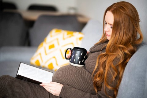 A pregnant woman with long red hair sits comfortably on a couch, reading a book and holding a mug.