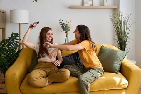 A happy lesbian couple shares a playful moment on a bright yellow sofa, surrounded by plants and