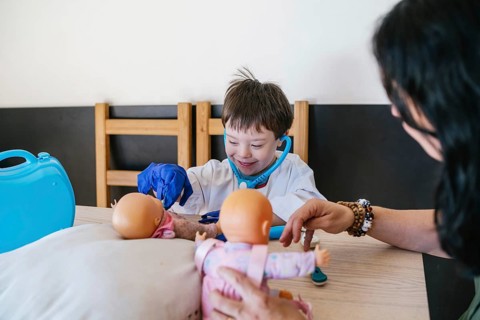A child with Down syndrome, dressed as a doctor, joyfully plays with toy dolls, using a stethoscope