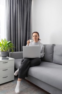 A woman work comfortably on a sofa with a laptop on her lap, sipping coffee. The scene includes