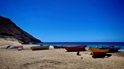 Colourful fishing boats lie on a sandy beach against a deep blue sea and clear sky backdrop,