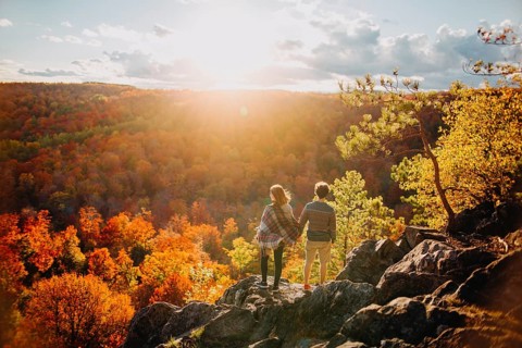 A couple stands on a rocky overlook, admiring the vibrant fall foliage under a bright autumn sky in