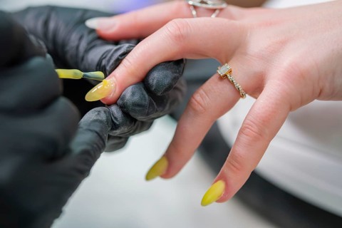 Close view of a nail technician in black gloves brushing glossy yellow polish onto a long almond