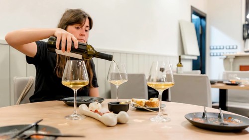 A woman pours white wine into glasses on a restaurant table set for dinner. The scene includes
