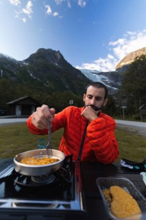 A traveler in a red jacket cooks a meal on a portable stove, set against the backdrop of majestic
