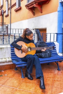 Vertical full length photo of an african american young woman playing guitar sitting on a bench