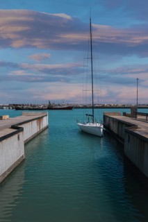 A sailboat calmly docked in a marina canal in Civitanova Marche, Italy. The sky is painted with