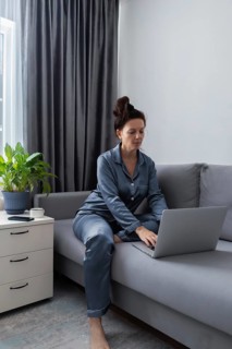A woman is focused on her laptop while sitting on a gray sofa, dressed in blue pajamas. Nearby, a