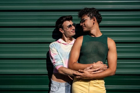A happy gay couple shares a tender embrace, with one partner holding the other from behind. They