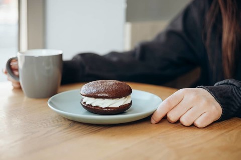 A delicious whoopie pie filled with cream rests on a plate, accompanied by a person enjoying coffee