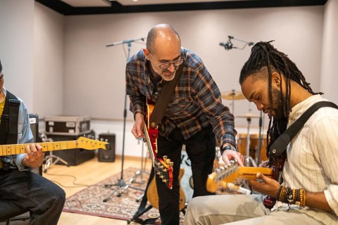 Musicians in a modern recording studio collaborate during a guitar session. The setting includes