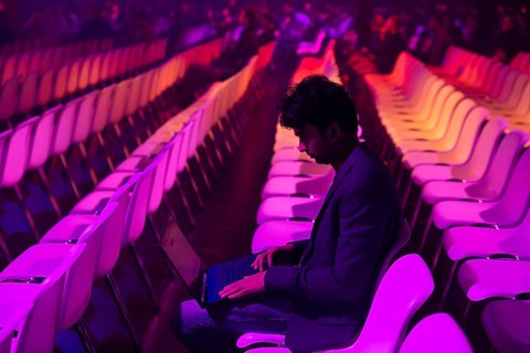 A indian man sits in a brightly lit auditorium with rows of colorful chairs at a conference of AI