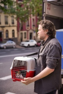A man carries a cat in a pet carrier on a bustling city street, heading to the veterinarian. The