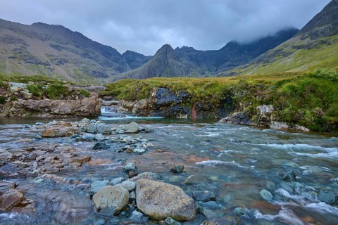 A clear river flows through a rocky landscape surrounded by high mountains under a cloudy sky,