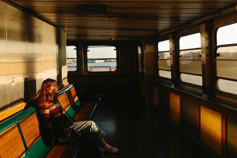 A woman sits alone on a ferry, absorbed in thought as the warm, golden light of the setting sun