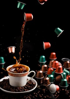 Coffee flows from a floating capsule into a cup, surrounded by capsules and coffee beans