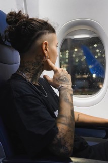 A young woman with elaborate tattoos looks through an airplane window, admiring the lights The