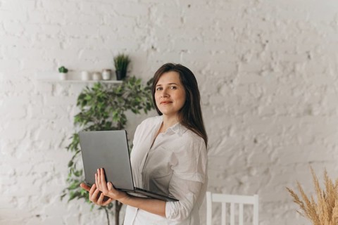 Nutritionist stands with a laptop in a modern office, wearing a white blouse. Background includes