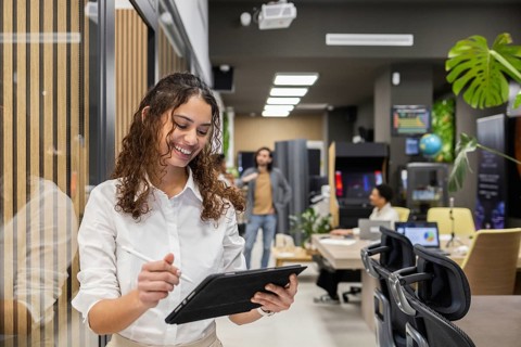 A cheerful professional in a modern office busily reviews information on a tablet. Colleagues work