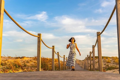 Woman wearing sunglasses and a patterned dress walking on a long wooden boardwalk, leading through