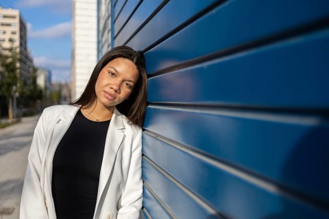 A confident woman in a modern, urban setting, dressed in business attire. She leans against a blue