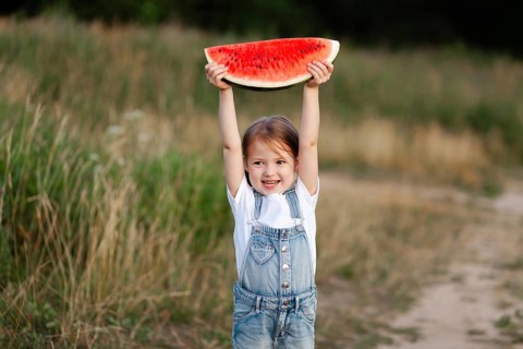A cheerful child holds a slice of watermelon above their head, enjoying a sunny day in nature. The