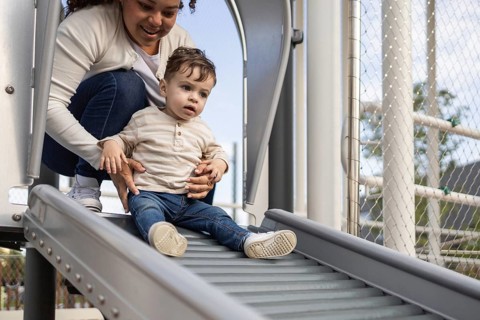 A mother and her baby enjoy a sunny day at the park. The mother helps her baby down a slide, both