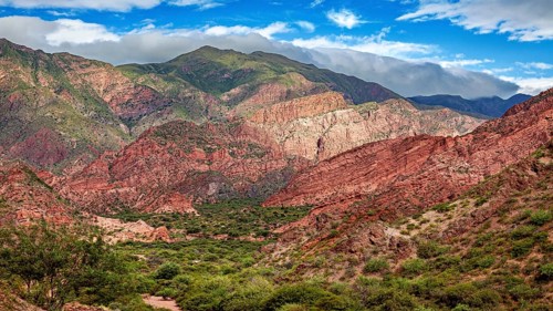 A mountainous landscape with red rocks and thick vegetation under a blue sky with clouds, The