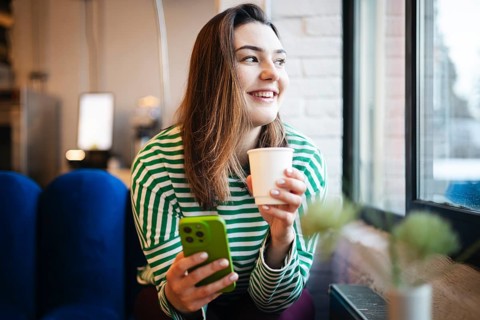 A woman in a striped shirt enjoys a coffee while using a smartphone at a cafe window, showcasing a