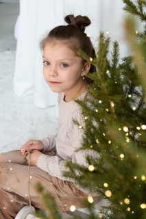 A young girl looks at the camera with soft eyes, seated beside a beautifully adorned Christmas tree