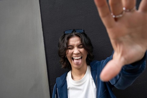 A person playfully sticks out their tongue and smiles broadly, wearing casual attire. Sunglasses