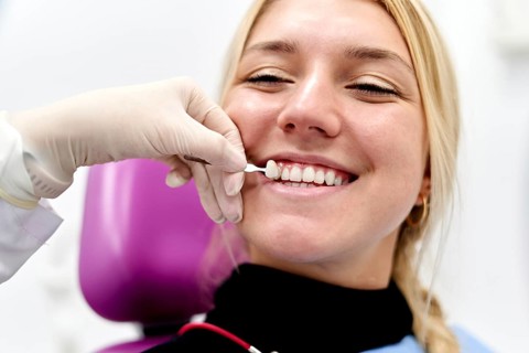 A dental professional in a clinic carefully matches tooth color for a smiling female patient,