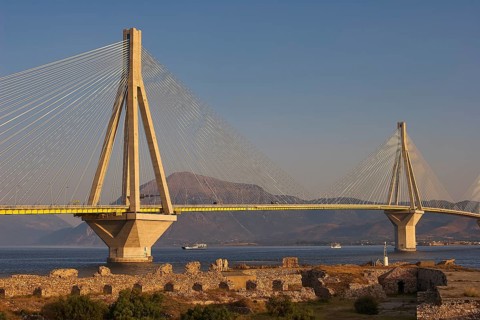 A long modern bridge stretches across the sea surrounded by ruins and mountains in the background,