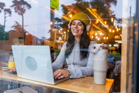 Woman wearing a beanie and headphones, smiling while working on her laptop at a cafe bar, enjoying